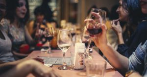 foto de mesa com taças de vinho em cima e pessoas em volta